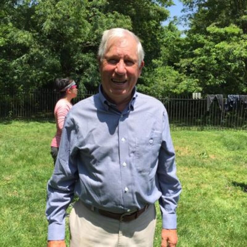 The image shows an older man standing in a grassy area, likely a backyard. He's wearing a blue button-down shirt and khaki pants. In the background, there's a woman and some trees. The lighting suggests it's a sunny day. The man is smiling, and the overall impression is casual and relaxed.